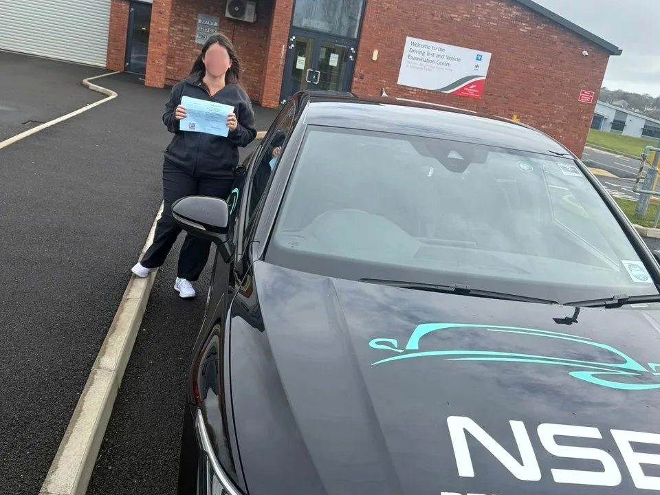 An anonymised learner holding a pass certificate beside the NSB Driver Training car at the Isle of Man test centre